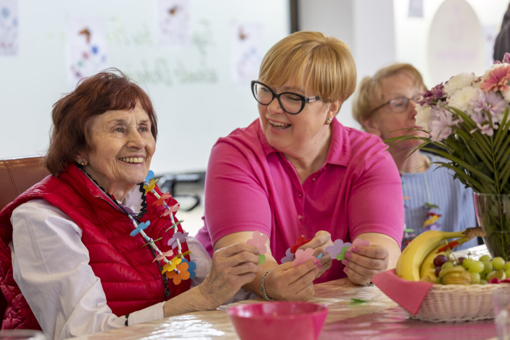 Seniorin und Pflegekraft basteln lächelnd am Tisch im Aufenthaltsraum – gemeinschaftliche Aktivierung.