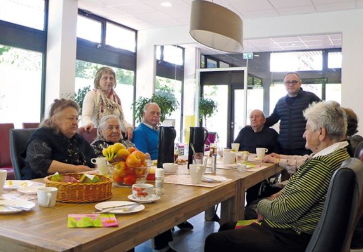 Seniorengruppe sitzt am großen Tisch im Speiseraum beim Frühstück; Geschirr und Obstschalen, helle Fensterfront.