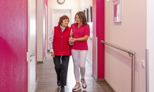 Pflegekraft begleitet Seniorin lächelnd durch hellen Flur mit Handläufen – sichere Orientierung und Unterstützung.
