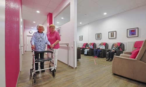 Seniorin mit Rollator im hellen Flur, von Pflegekraft begleitet; rechts Gäste im Aufenthaltsbereich mit Sesseln.