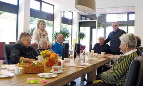 Seniorengruppe sitzt am großen Tisch im Speiseraum beim Frühstück; Geschirr und Obstschalen, helle Fensterfront.